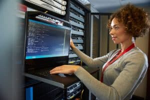 A woman with curly hair is working on a laptop in a server room. Wearing a gray sweater and a red lanyard, she is deeply focused on the VCSO code displayed on the screen. Server racks filled with equipment are visible in the background.
.
