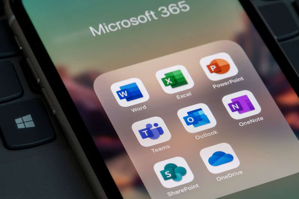 Close-up of a smartphone screen displaying Microsoft 365 app icons, including Word, Excel, PowerPoint, Teams, Outlook, OneNote, SharePoint, and OneDrive. A blurred laptop keyboard is in the background.