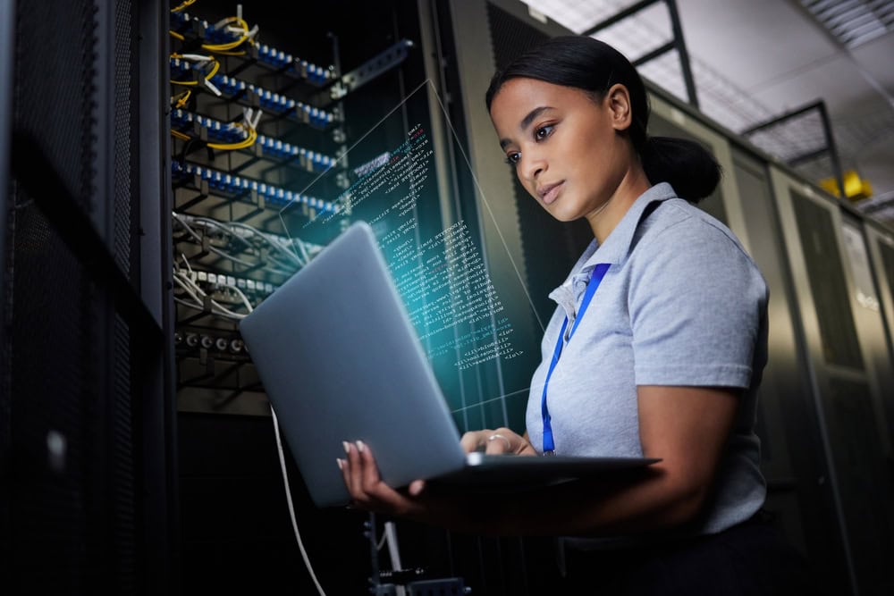 A woman in a server room works on a laptop. She is wearing a gray polo shirt and has badges around her neck. Digital code is visible on the image, suggesting programming or data analysis. Server racks are visible in the background.