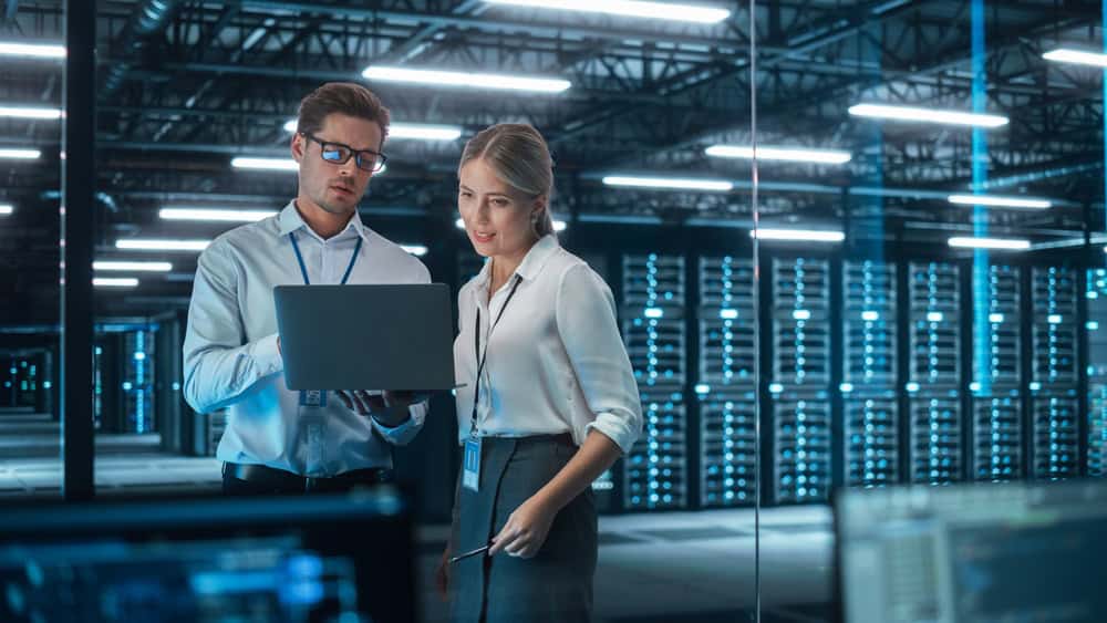 Two professionals stand in a modern server room, looking at a laptop. Rows of server racks with blue lights are in the background, and both people wear ID badges and business attire.