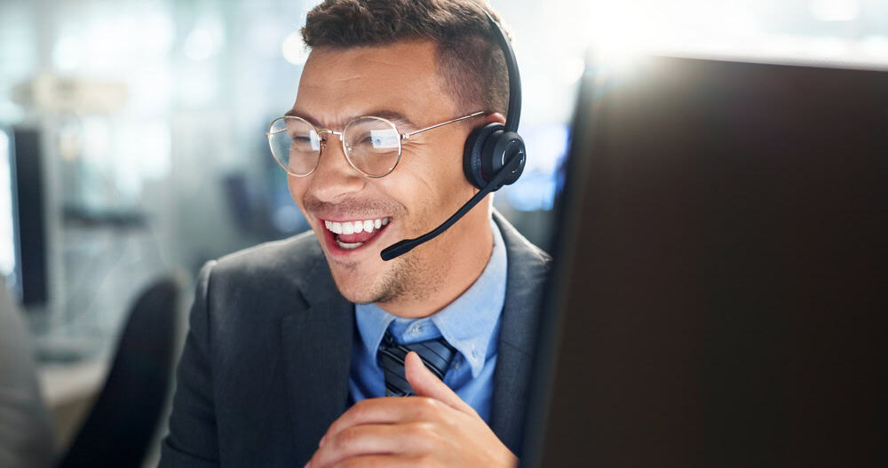 A smiling man wearing glasses and a headset sits at a desk, likely working in a call center or customer service, with a computer monitor in front of him.