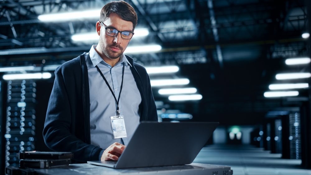 A man wearing glasses and a smart casual outfit stands in a dimly lit data center, focusing on his laptop. He has an ID badge around his neck, with server racks in the background. The environment is modern and high-tech.