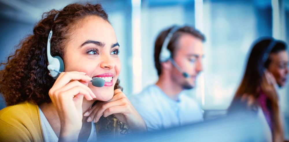 A smiling woman in a headset works in a cybersecurity & IT support call center, with two colleagues beside her in a professional setting.