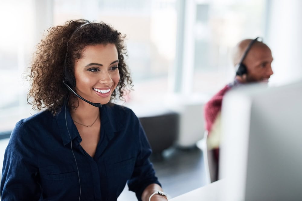 A woman with curly hair is smiling and wearing a headset while working at a computer. She is dressed in a dark blue shirt. In the background, a man with a shaved head, also wearing a headset, is slightly out of focus.