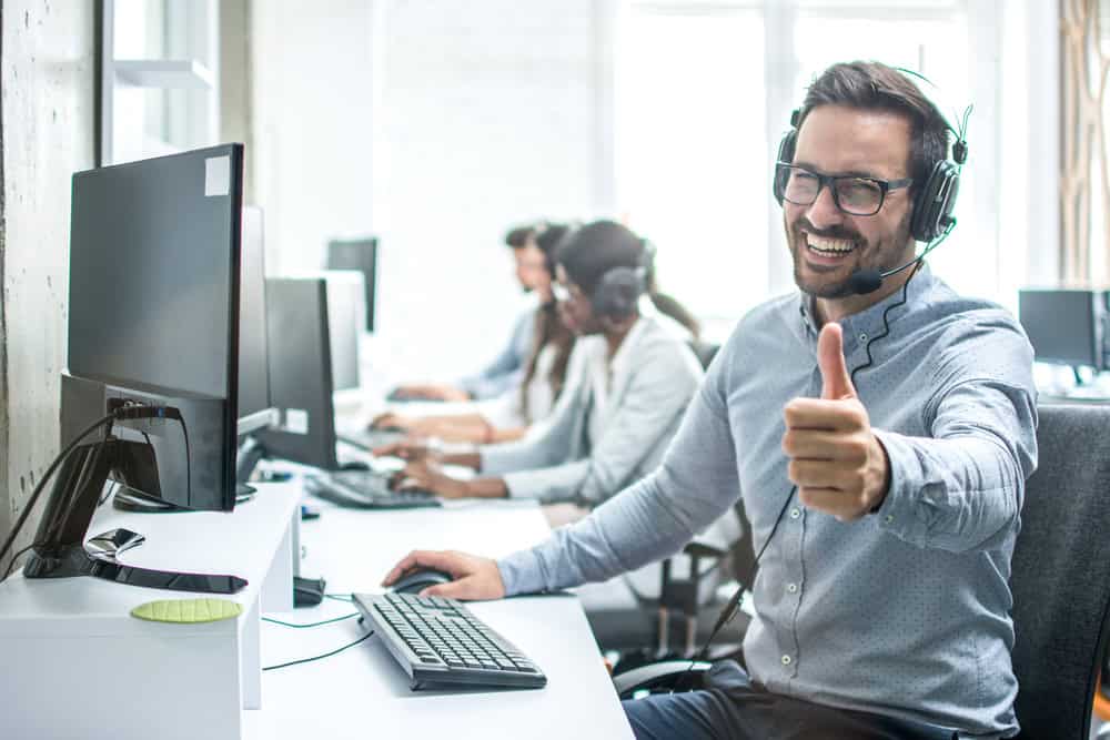 A man in a headset sits at a desk, smiling and giving a thumbs-up while providing IT support services. Colleagues work in the background.