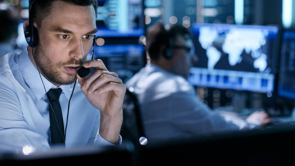 A man in a headset focuses on his screen in a busy IT support room, managing Cloud Services for Business, with maps on monitors.