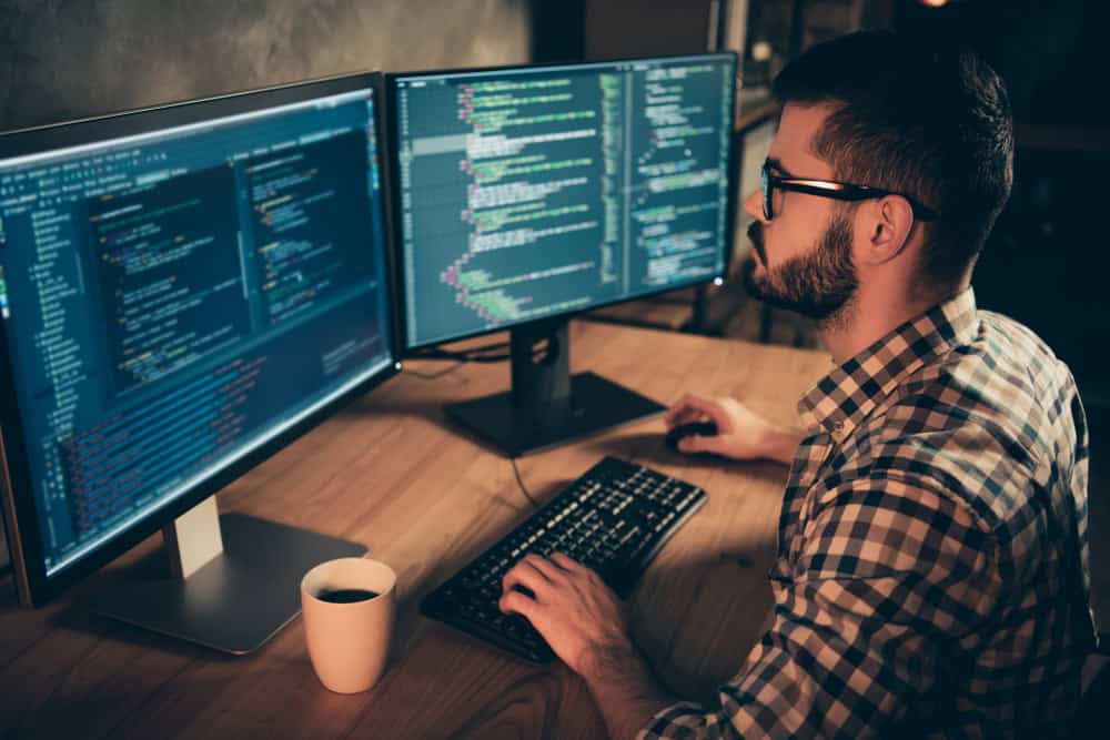 A person wearing glasses and a plaid shirt is typing on a keyboard while looking at code on two monitors. A cup is placed on the wooden desk next to the keyboard. The room has a dimly lit ambiance.