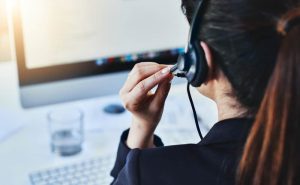 A person wearing a headset sits at a desk with a computer monitor in the background. They are holding the microphone of the headset near their mouth. The setting appears to be an office environment, with a glass of water on the desk.