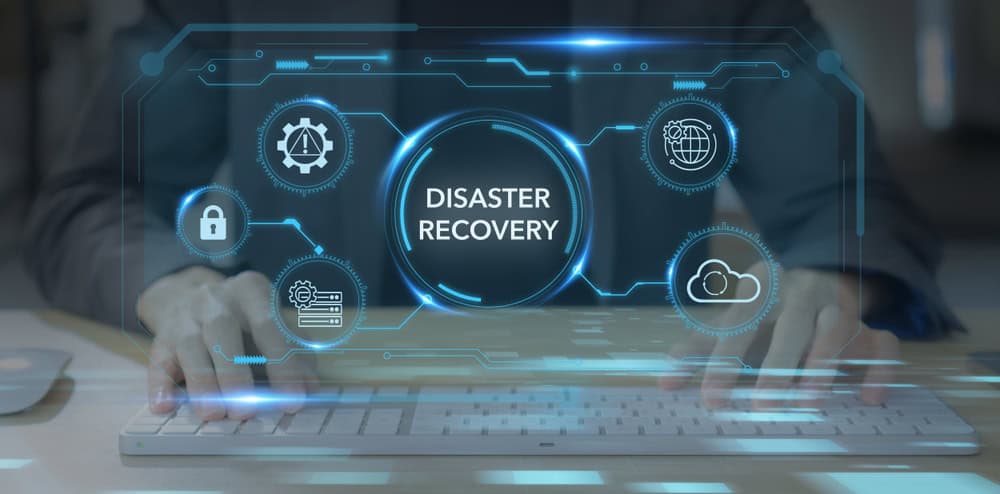 ALT text: Person typing on a keyboard with a virtual interface showing icons and the words "Disaster Recovery" in the center. Icons include a lock, cloud, globe, and gears. Blue digital graphics overlay the scene.