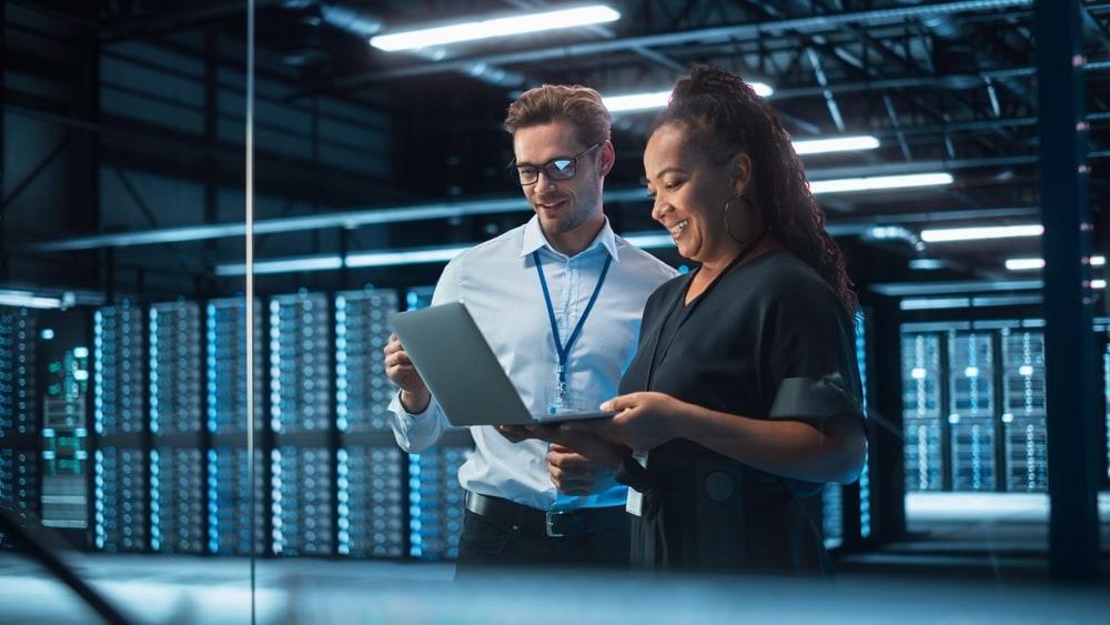 Two people stand in a modern data center. One holds a laptop, and they both appear to be examining information. Behind them are rows of server racks, illuminated by blue lights.