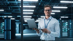 A smiling person wearing glasses and a white shirt stands in a modern data center, holding an open laptop. The background features illuminated servers and industrial lighting, highlighting the cutting-edge cybersecurity measures safeguarding vital information.