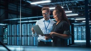 Two people stand in a modern data center. One holds a laptop, and they both appear to be examining information. Behind them are rows of server racks, illuminated by blue lights.
