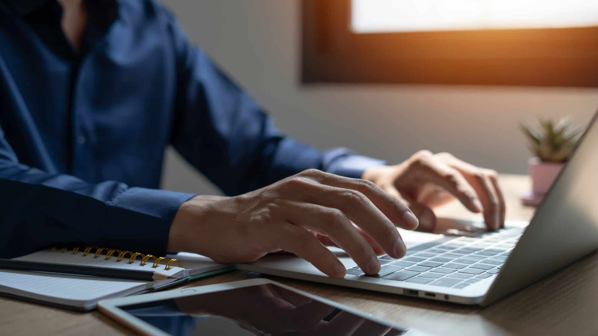 A person in a dark blue shirt types on a laptop at a wooden desk, with a notebook, pen, and smartphone nearby. Sunlight shines through a window in the background, creating a warm atmosphere.