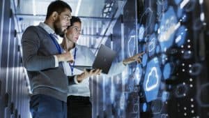 Two IT professionals stand in a server room, examining a large transparent screen with digital icons, including a cloud symbol. One holds a laptop while the other points at the display, both focused on the data.