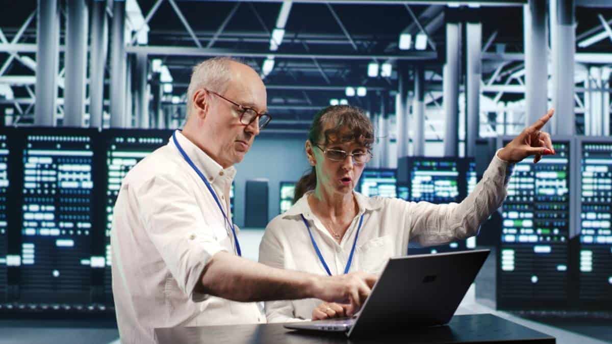 Two people wearing glasses and white shirts work together on a laptop in a server room. The woman is pointing forward while the man types on the laptop. Rows of server racks are visible in the background.
