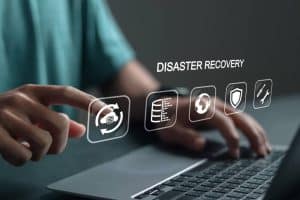 A person uses a laptop, touching virtual icons related to disaster recovery, such as a database, files, globe, shield, and tools, with the words "DISASTER RECOVERY" floating above.