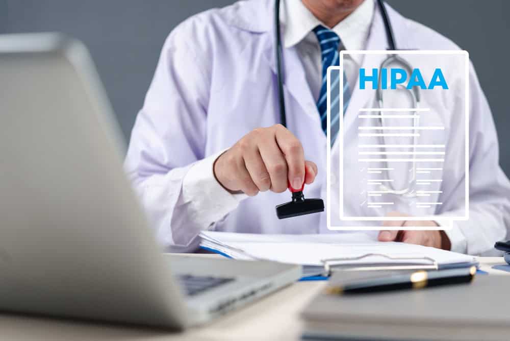 A doctor in a white coat uses a stamp on paperwork at a desk with a laptop. A transparent digital graphic displays “HIPAA” and a lock icon, symbolizing healthcare data privacy and security compliance.