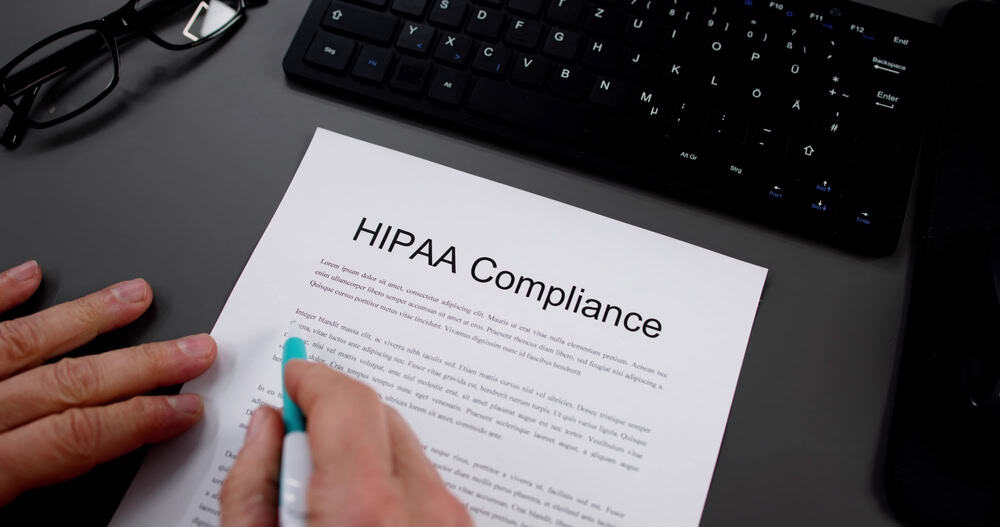 A person highlights text on a printed document titled “HIPAA Compliance,” with a keyboard, glasses, and pen visible on a gray desk.