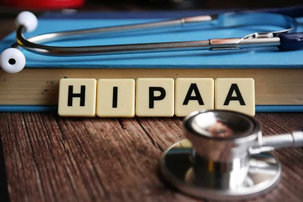 Scrabble tiles spelling "HIPAA" rest on a closed book, with a stethoscope and another medical instrument nearby, symbolizing healthcare privacy and compliance.
