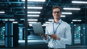A smiling man wearing glasses and a lanyard holds an open laptop in a brightly lit data center filled with server racks at an IT company serving Illinois and Indiana.