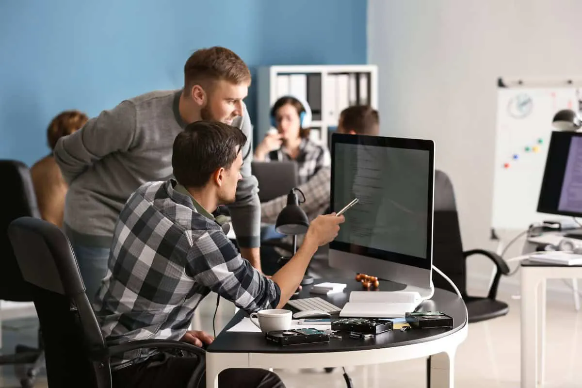 Two men working at a desk with a computer monitor in a modern office at an IT company in Illinois and Indiana, one seated and pointing at the screen while the other stands beside him. Other coworkers work at desks in the background.